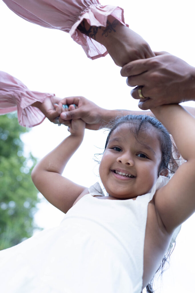 The hands of mother, father, and young daughter holding each other while girl smiles at the camera